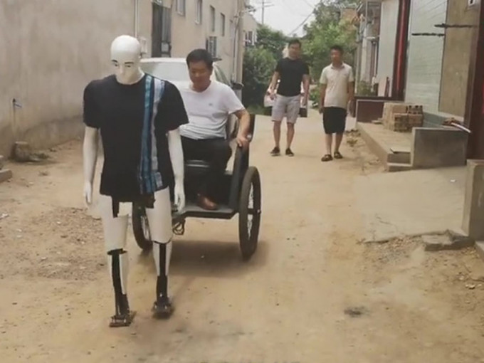 段男坐自己發明的拉車，由車夫機械人拉着在村内行走。（網圖）