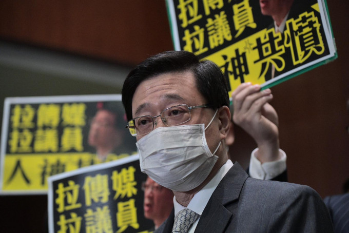 李家超在立法會見傳媒時，民主派議員舉示威標語。