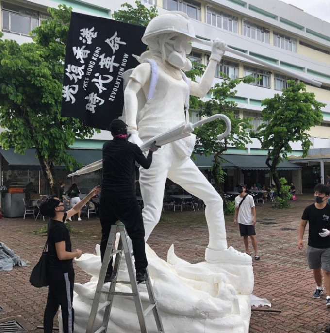 雕像戴有头盔、眼罩及「猪嘴」口罩，手持雨伞，以及写有「光复香港、时代革命」旗帜。连登图片