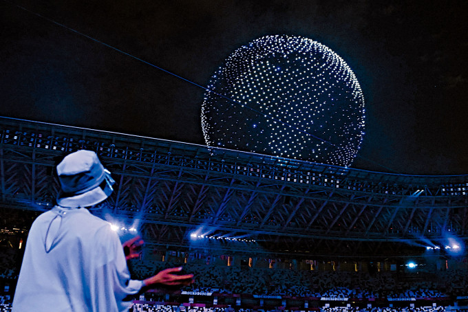■選手和義工上周五在東奧開幕禮，觀看無人機燈光騷。多架無人機在夜空中組成象徵和平的藍色地球影像。