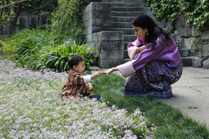 小王子坐在皇宮花園草地上，伸手觸碰媽媽佩瑪的手。