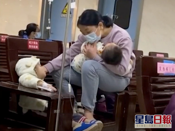 年轻妈妈一手抱著熟睡的幼子,另一手轻轻扶著吊盐水的大儿子。网图