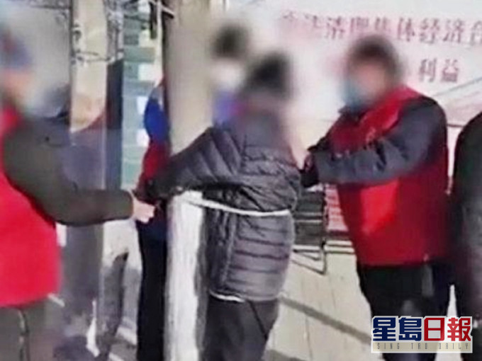 一名身穿黑色羽絨的老人,被一群穿紅色背心外套的人綁在樹上,並遭到辱罵。影片截圖