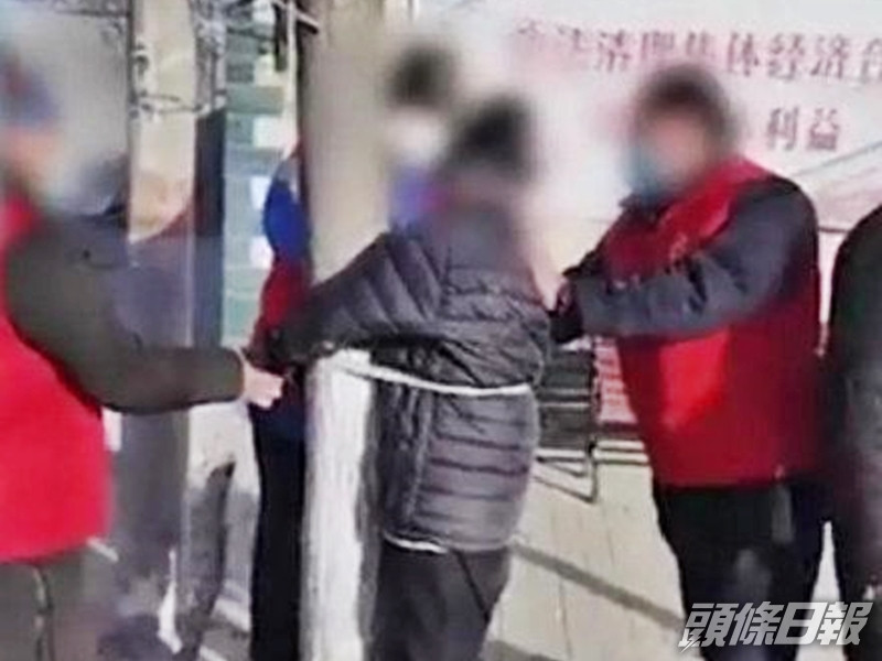 一名身穿黑色羽絨的老人,被一群穿紅色背心外套的人綁在樹上,並遭到辱罵。影片截圖